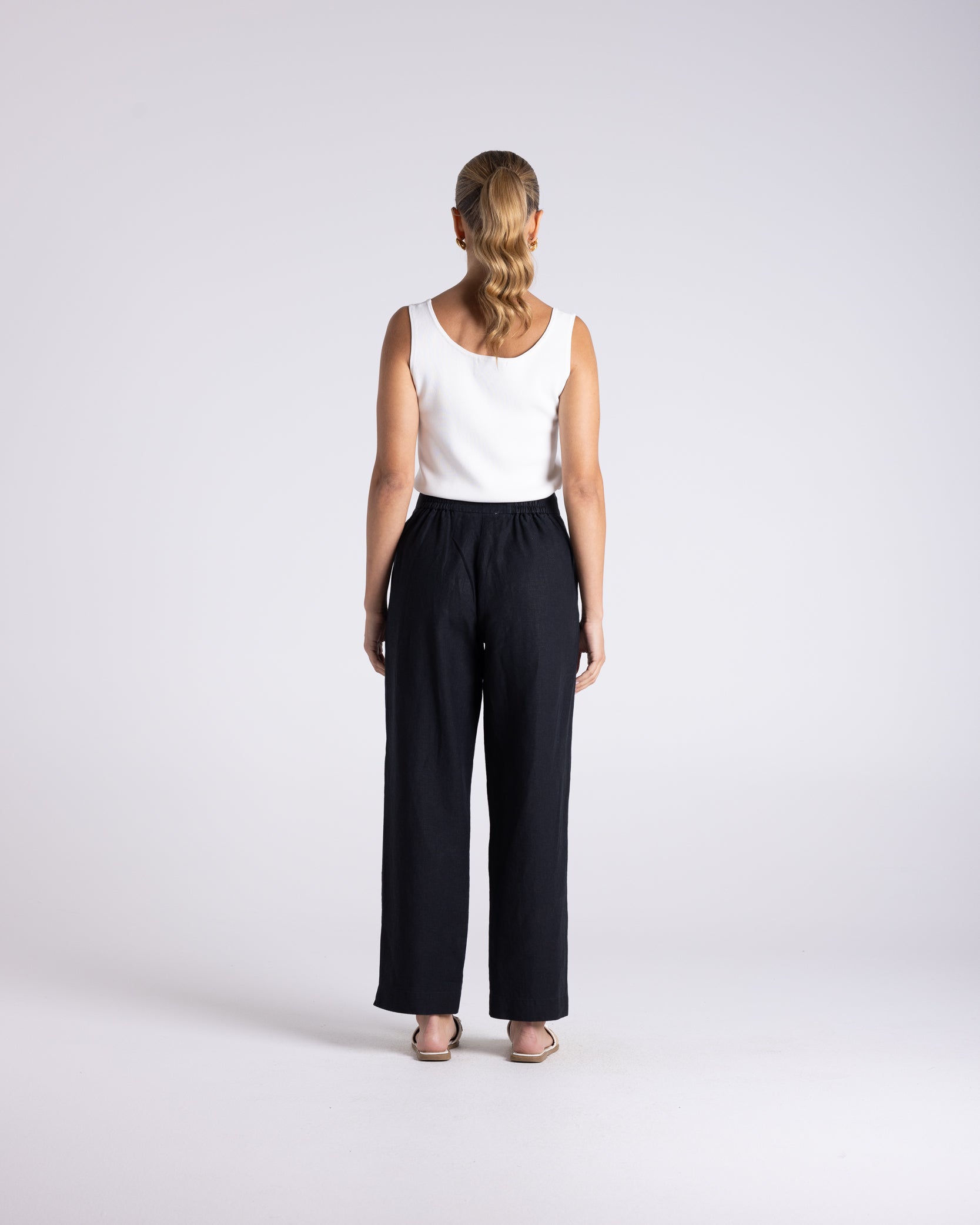 A woman standing with her back to the camera, wearing a white sleeveless top and black wide leg trousers against a light gray studio background.