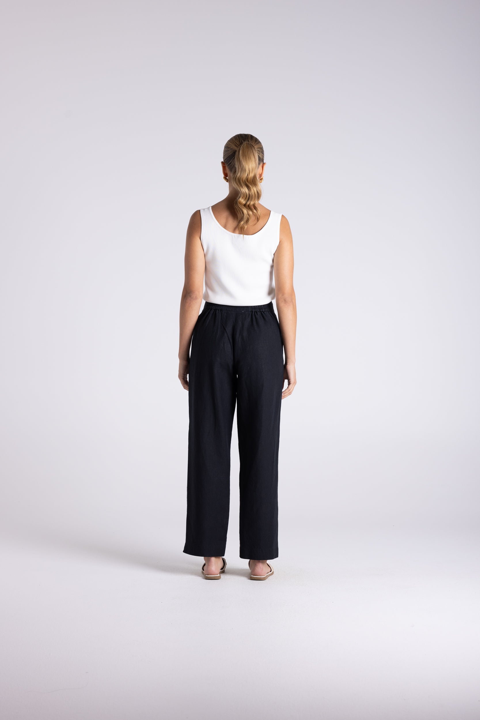 A woman standing with her back to the camera, wearing a white sleeveless top and black wide-leg trousers against a light gray studio background.