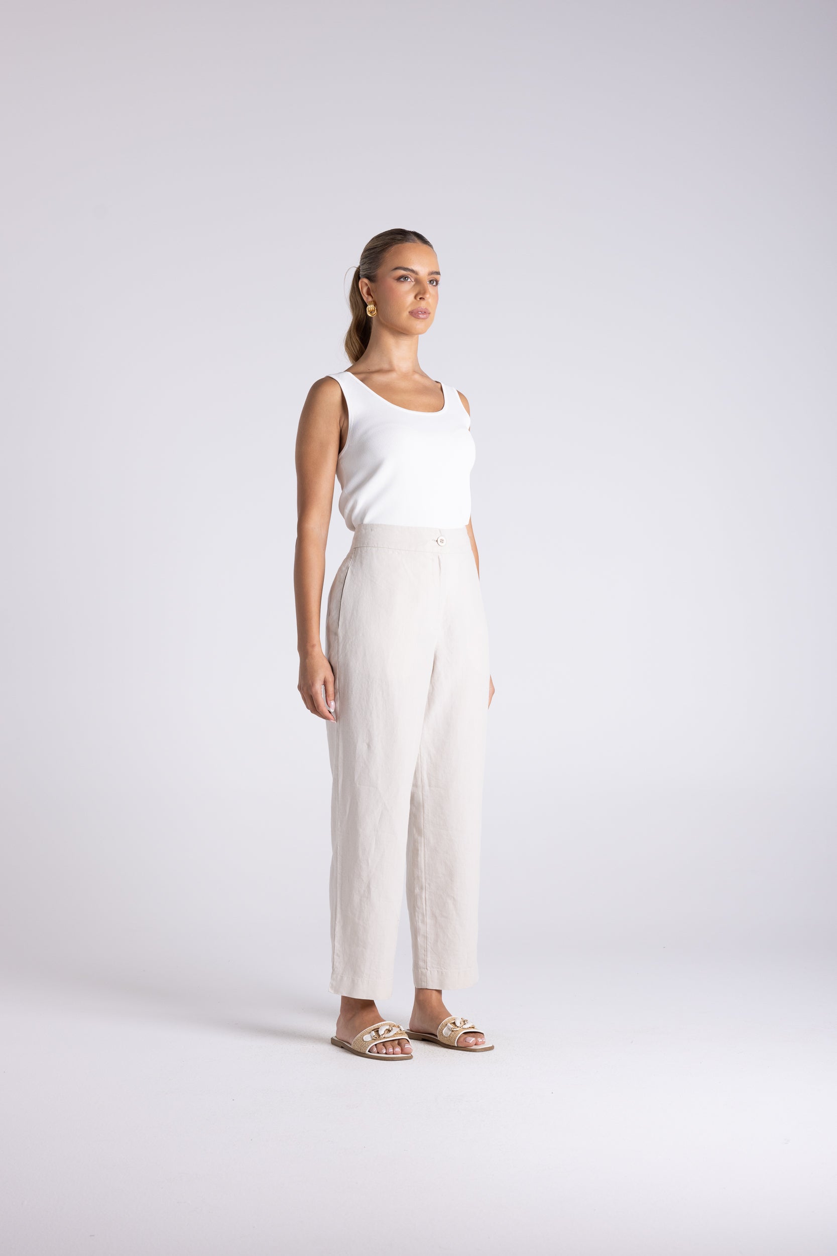 A woman stands in a studio wearing a white sleeveless top and off-white wide-leg trousers with beige sandals, photographed against a light gray background.