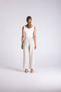 A woman stands with her back to the camera wearing a white sleeveless top and light beige wide-leg trousers, photographed against a seamless light gray studio background.