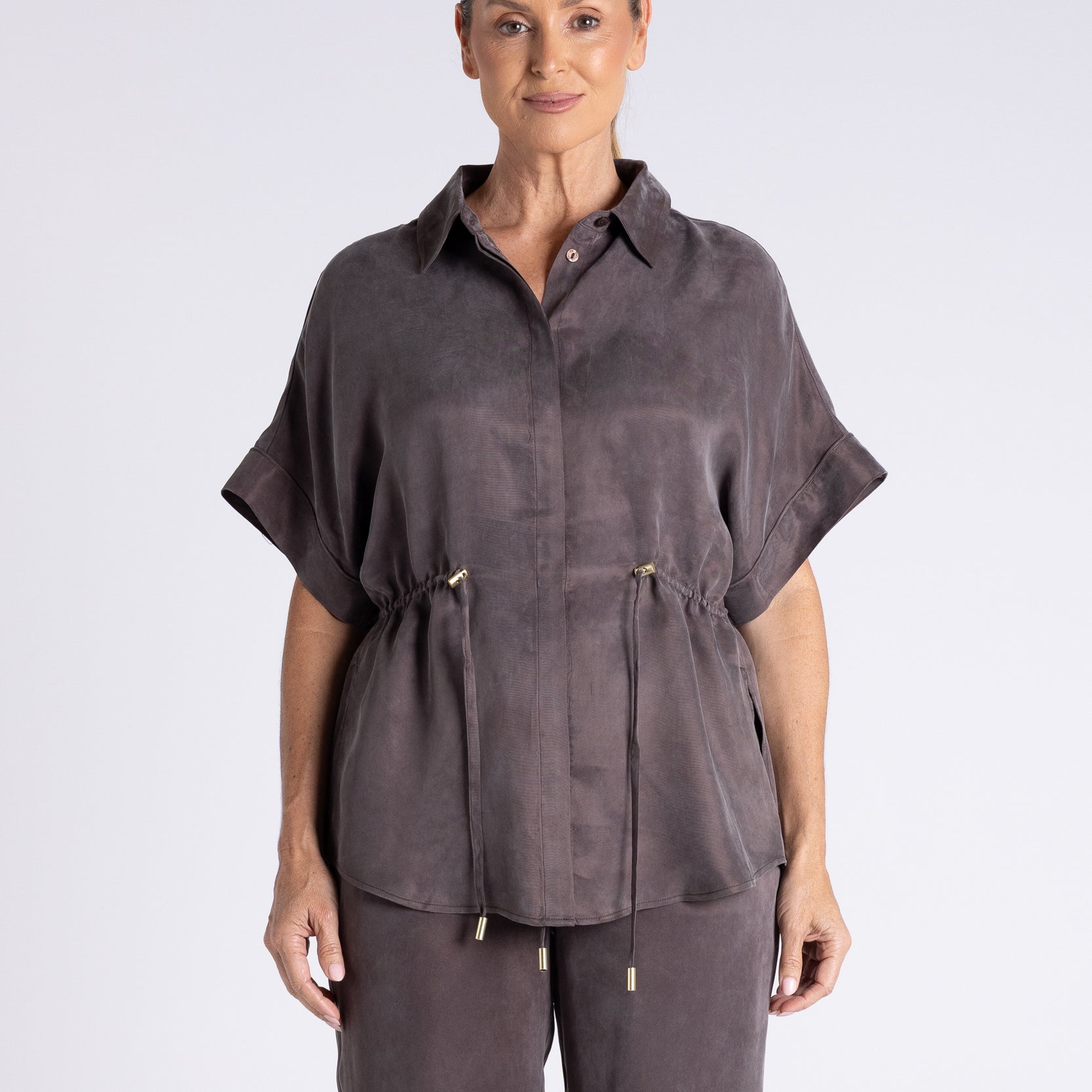 A woman modeling a dark gray short-sleeve button-up shirt with a drawstring waist, paired with matching drawstring trousers, against a light gray studio background.