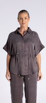 A woman modeling a dark gray short-sleeve button up shirt with a drawstring waist, paired with matching drawstring trousers, against a light grey studio background.