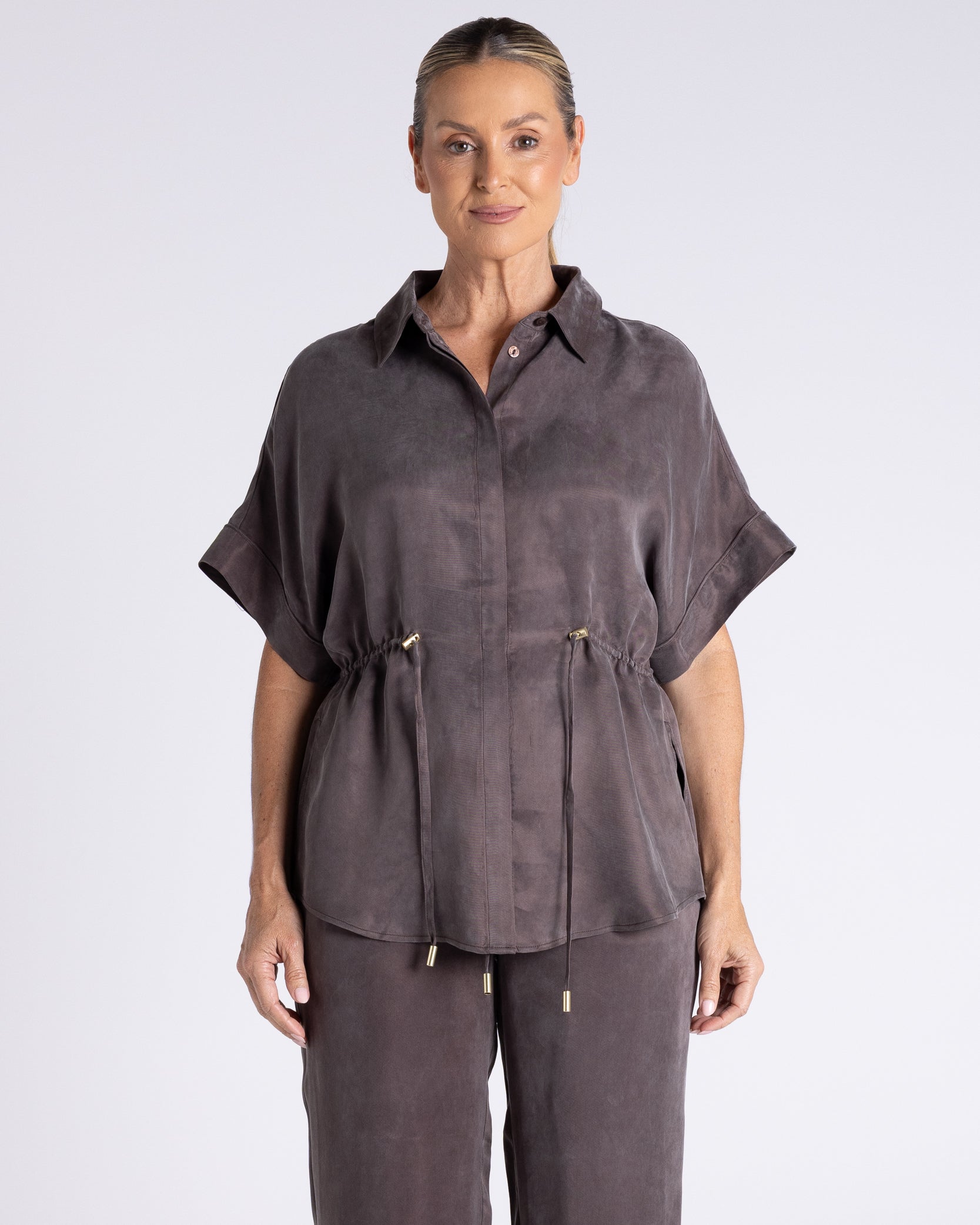 A woman modeling a dark gray short-sleeve button-up shirt with a drawstring waist, paired with matching drawstring trousers, against a light gray studio background.