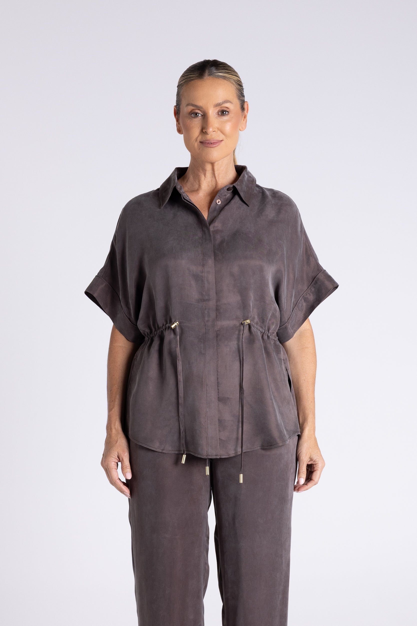 A woman modeling a dark gray short-sleeve button-up shirt with a drawstring waist, paired with matching drawstring trousers, against a light gray studio background.