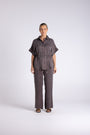 A woman stands against a light gray studio background wearing a matching dark gray short-sleeve button-up shirt and wide-leg trousers set.