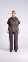 A woman stands against a light gray studio background wearing a matching dark gray short sleeve button up shirt and wide leg trousers set.