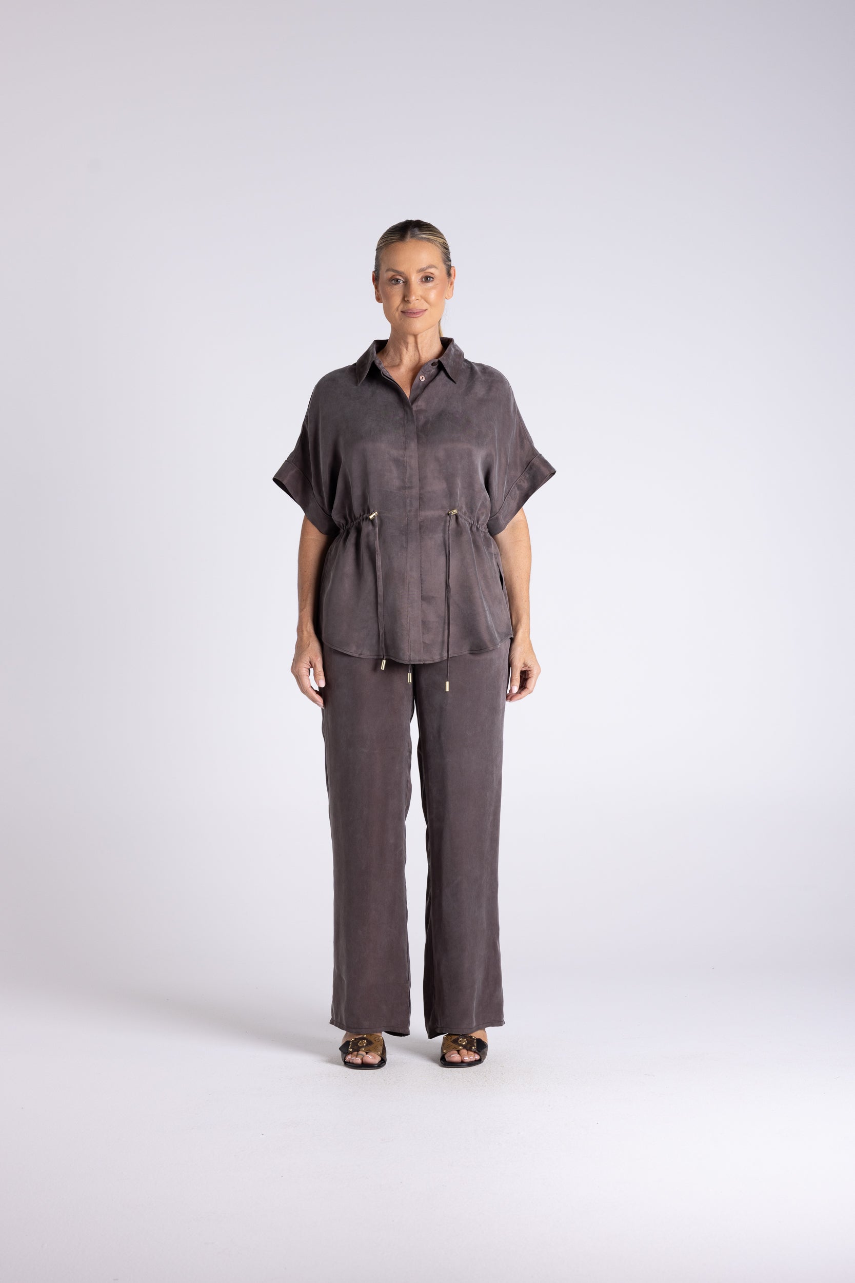 A woman stands against a light gray studio background wearing a matching dark gray short-sleeve button-up shirt and wide-leg trousers set.