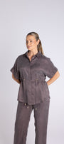 A woman modeling a matching two-piece lounge set in a dark greyish brown suede like fabric, featuring a short sleeve button up shirt and matching drawstring pants, against a light grey studio background.