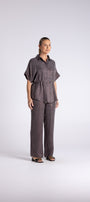 A woman modeling a dark gray two piece linen set consisting of a short sleeve button up shirt and matching wide leg trousers, standing against a light grey studio background.