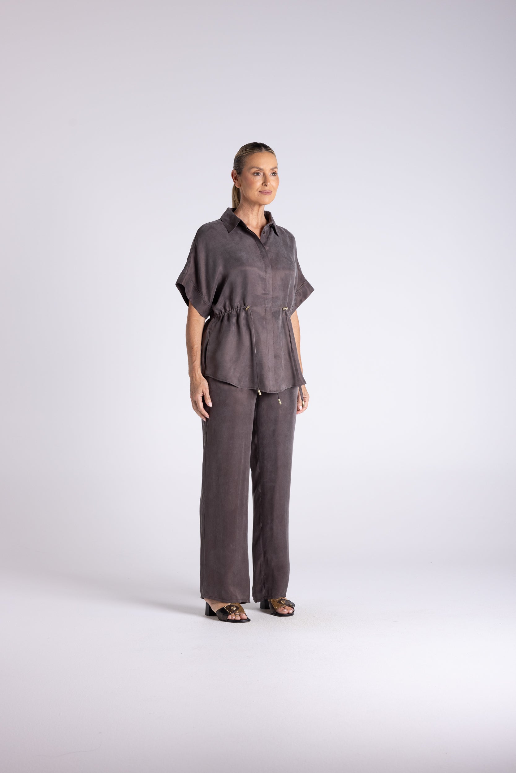 A woman modeling a dark gray two-piece linen set consisting of a short-sleeve button-up shirt and matching wide-leg trousers, standing against a light gray studio background.