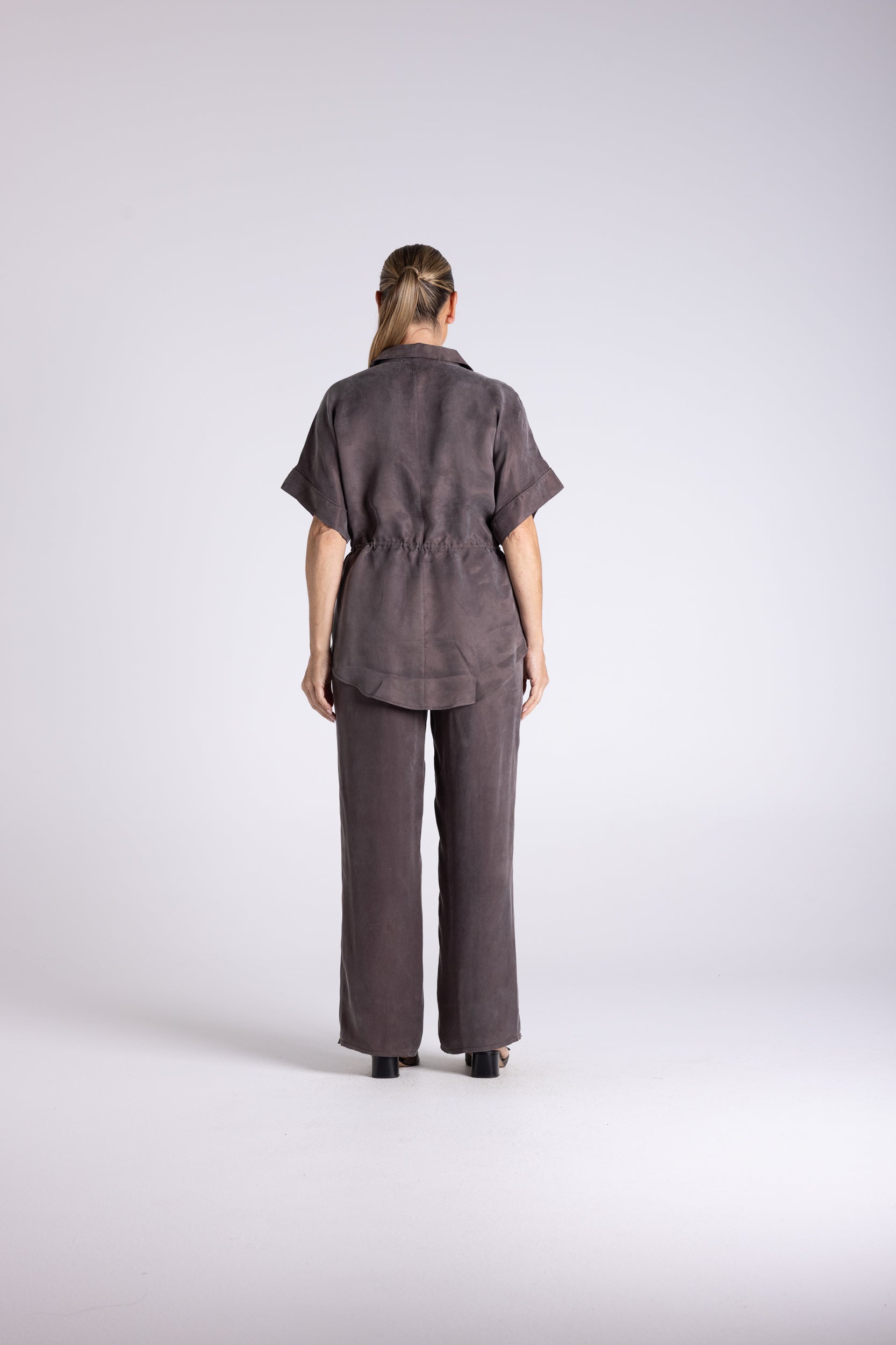 A model viewed from the back wearing a dark brown short-sleeve collared shirt and matching wide-leg trousers against a light gray studio background.