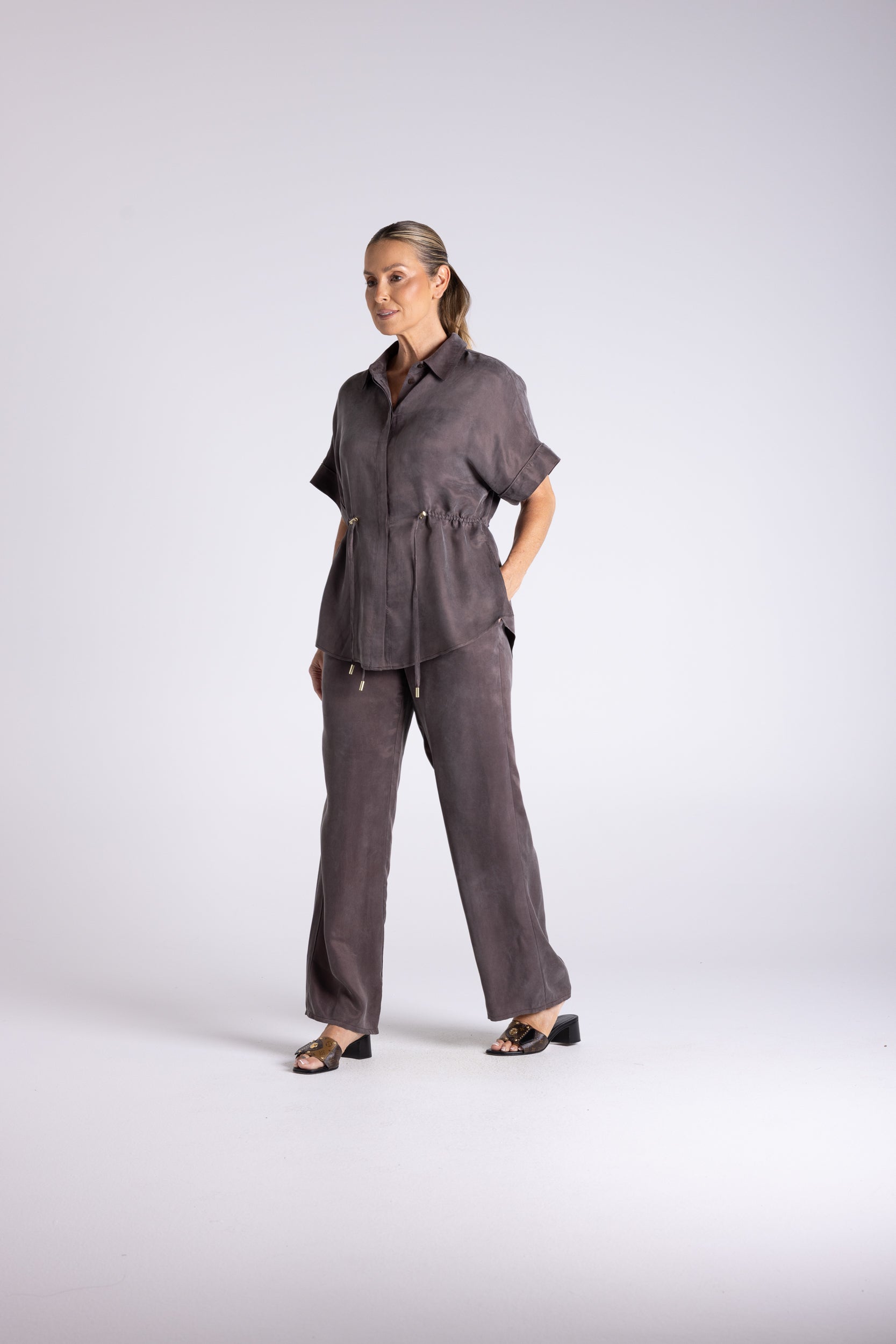 A woman modeling a dark gray short-sleeve button-up shirt and matching wide-leg trousers, standing against a light gray studio background.