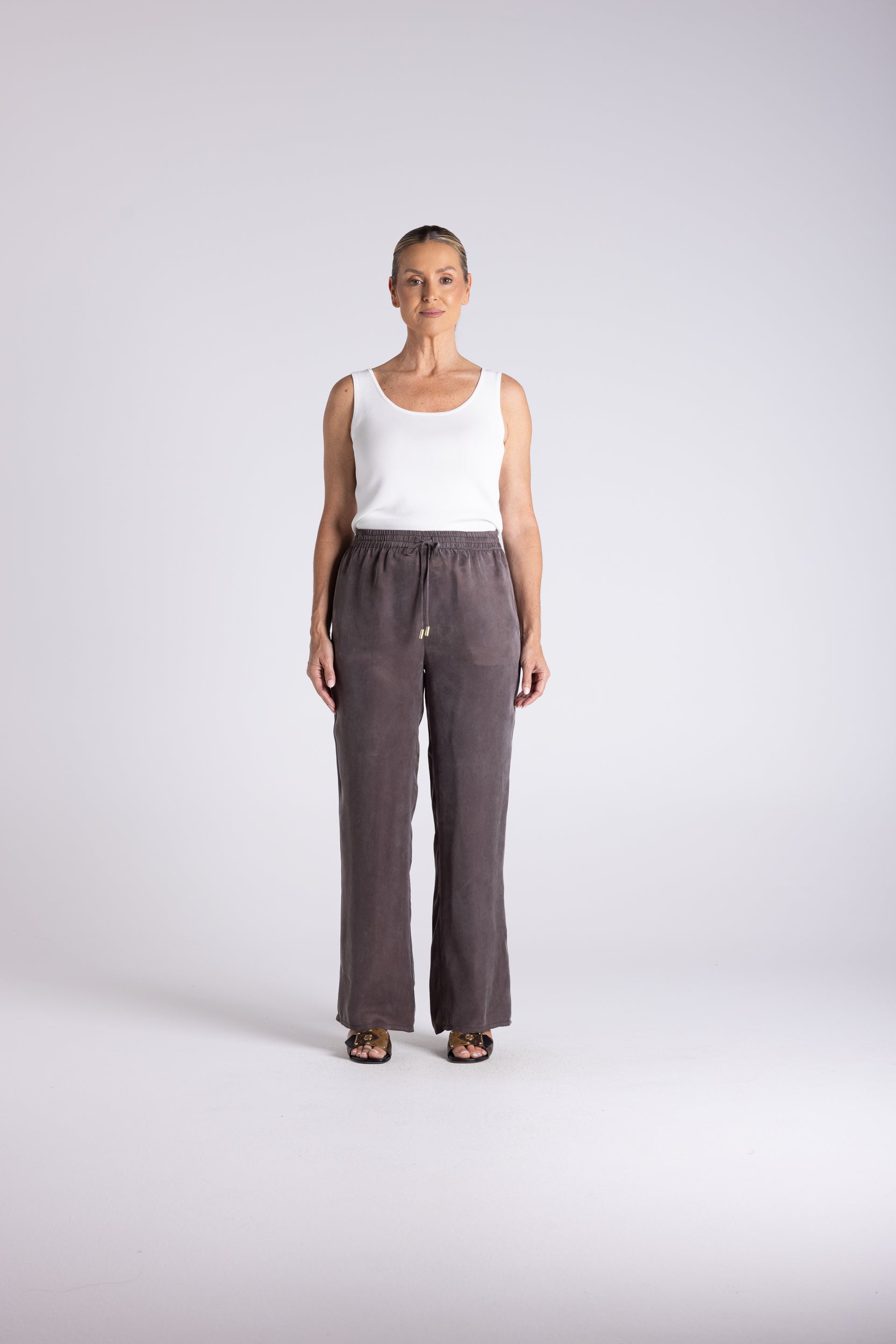 A woman stands against a light gray studio background wearing loose, wide-leg taupe-brown pants with a white sleeveless top and dark sandals.
