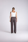 A woman viewed from behind wearing a white sleeveless top and dark brown wide-leg trousers, standing against a light gray studio background.