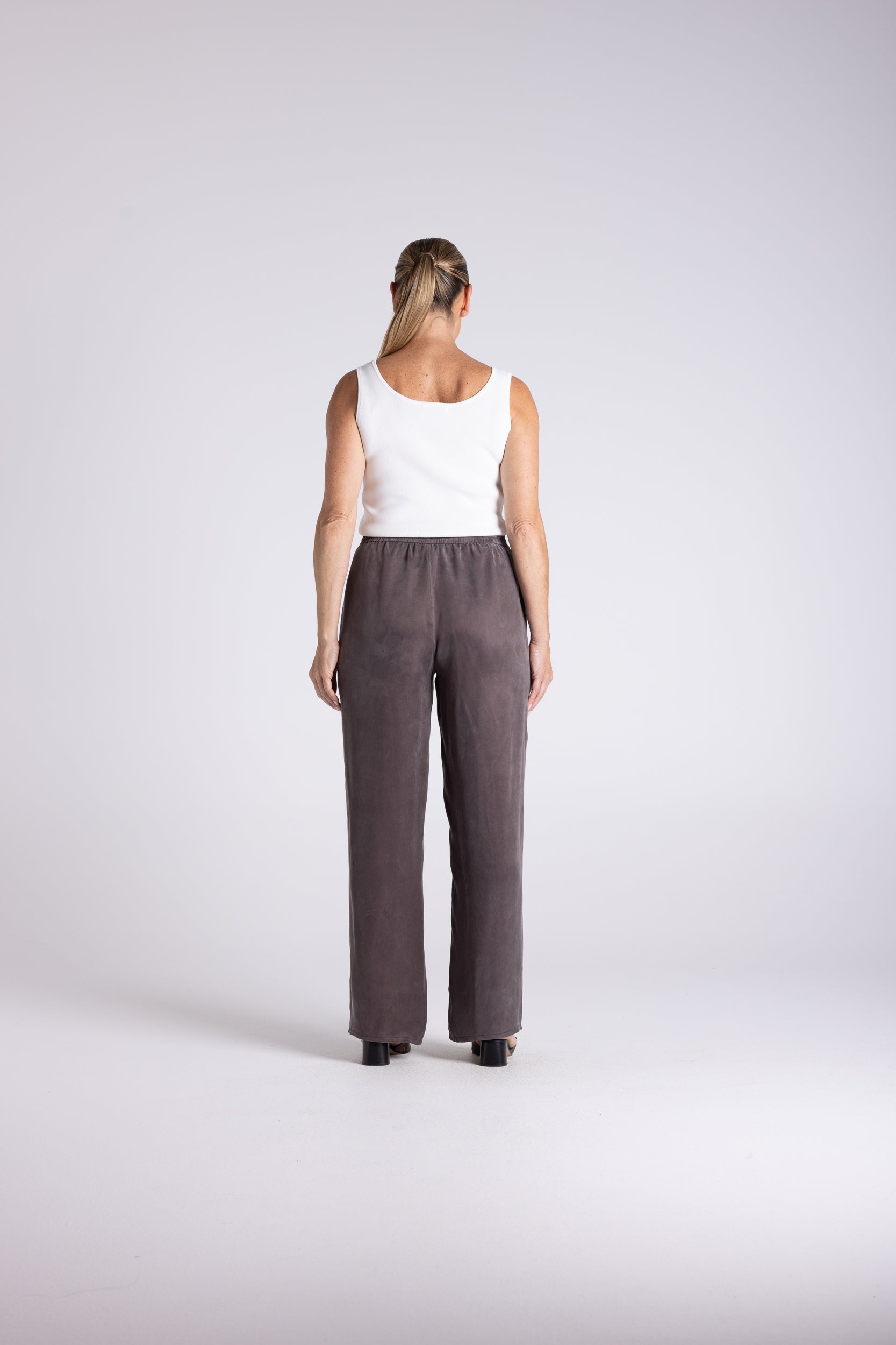 A woman viewed from behind wearing a white sleeveless top and dark brown wide-leg trousers, standing against a light gray studio background.