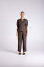 A model stands against a light gray studio background wearing a matching dark brown two-piece outfit consisting of a short-sleeve top with a split neckline and wide-leg trousers.