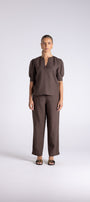 A model stands against a light gray studio background wearing a matching dark brown two-piece outfit consisting of a short-sleeve top with a split neckline and wide-leg trousers.