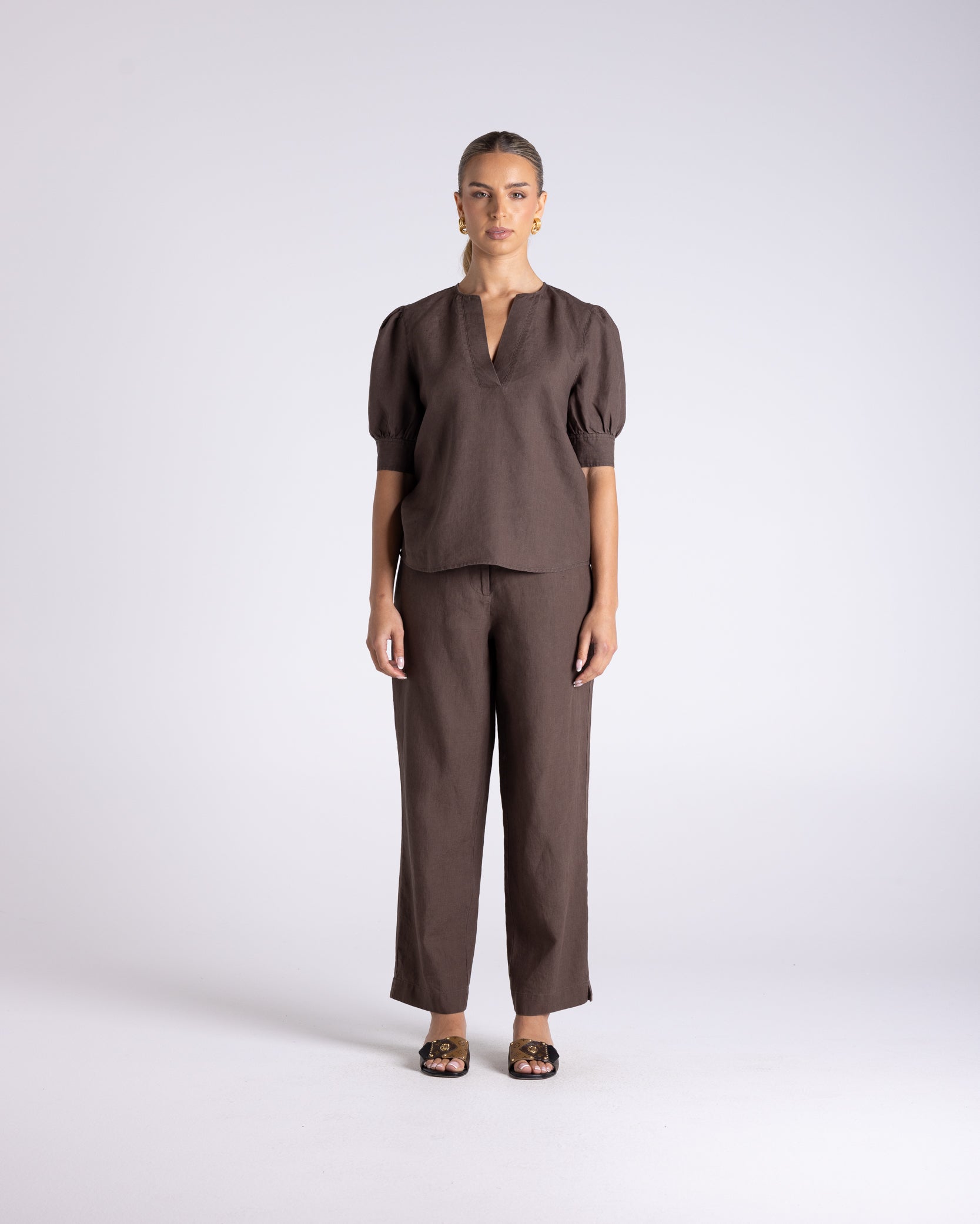 A model stands against a light gray studio background wearing a matching dark brown two-piece outfit consisting of a short-sleeve top with a split neckline and wide-leg trousers.