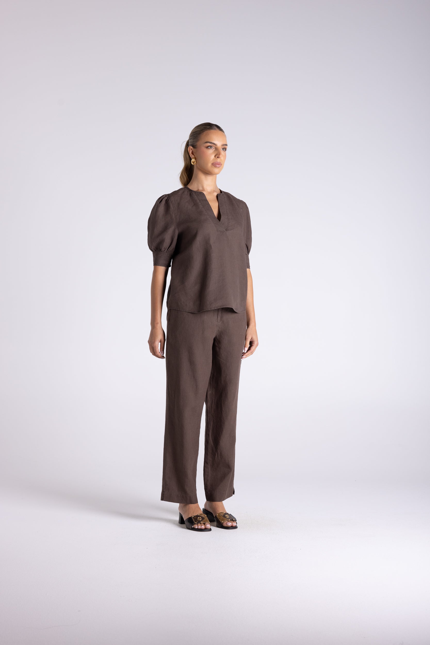 A woman modeling a matching dark brown two-piece set consisting of a short-sleeve cropped top with puff sleeves and wide-leg trousers, photographed against a light gray studio background.