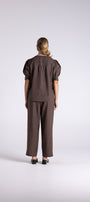 A model viewed from the back wearing a dark brown short-sleeve top with puffed shoulders and matching wide-leg trousers, standing against a light gray studio background.