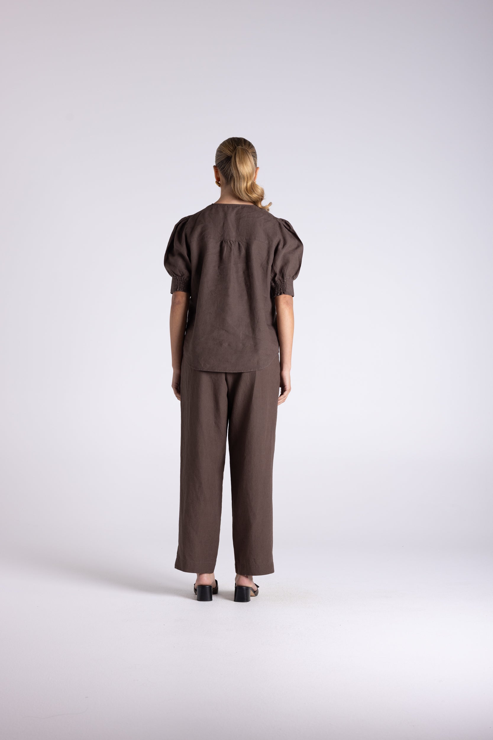 A model viewed from the back wearing a dark brown short-sleeve top with puffed shoulders and matching wide-leg trousers, standing against a light gray studio background.