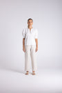 A woman stands against a light gray studio background wearing a white short-sleeve blouse with puffed shoulders and a split neckline, paired with wide-leg light beige trousers and beige sandals.