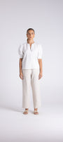 A woman stands against a light gray studio background wearing a white short-sleeve blouse with puffed shoulders and a split neckline, paired with wide-leg light beige trousers and beige sandals.
