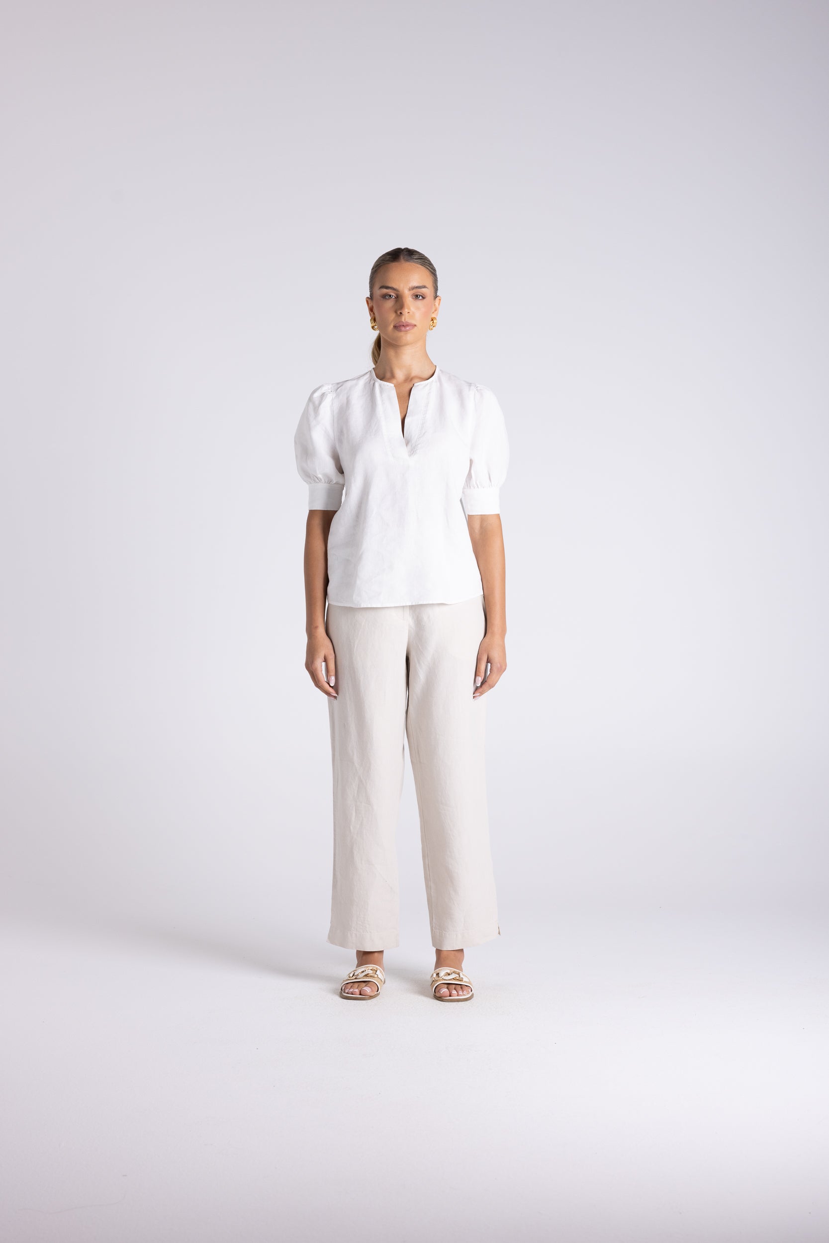 A woman stands against a light gray studio background wearing a white short-sleeve blouse with puffed shoulders and a split neckline, paired with wide-leg light beige trousers and beige sandals.