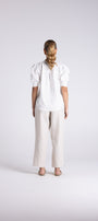A woman viewed from behind wearing a white puff-sleeve blouse and light beige wide-leg trousers, standing against a light gray studio background.
