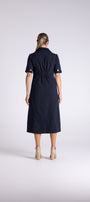 A woman viewed from behind wearing a dark navy midi dress with short sleeves, a collar, and a gathered waist, paired with beige ankle boots, against a light gray studio background.