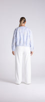 A model shown from the back wearing a light blue patterned long-sleeve top and white wide-leg trousers, standing against a light gray studio background.