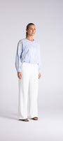 A woman modeling a light blue patterned long-sleeve blouse and white wide-leg trousers against a light gray studio background.