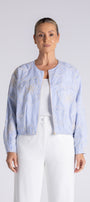 A woman modeling a light blue cropped jacket with white embroidery, worn over a white top and white trousers, against a light gray studio background.