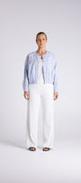 A model stands against a light gray studio background wearing a light blue cropped denim jacket over a white top and white wide-leg trousers.