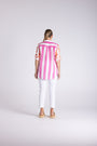 A woman viewed from behind wearing a pink and white vertically striped short-sleeve button-up shirt, white cropped pants, and tan sandals, standing against a light gray studio background.