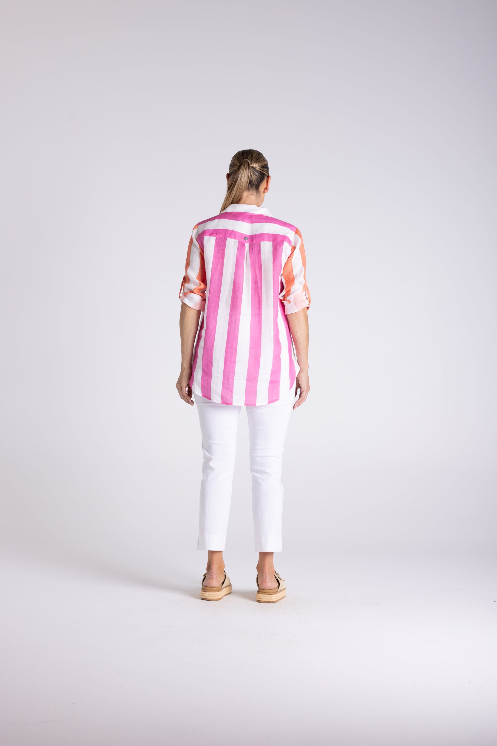 A woman viewed from behind wearing a pink and white vertically striped short-sleeve button-up shirt, white cropped pants, and tan sandals, standing against a light gray studio background.