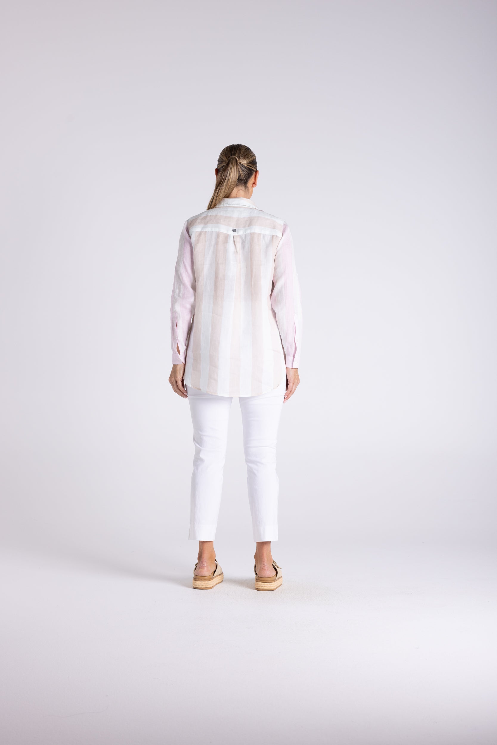 A woman viewed from behind wearing a white long-sleeve button-up shirt with a sheer yoke, paired with white cropped pants and tan sandals, against a light gray studio background.