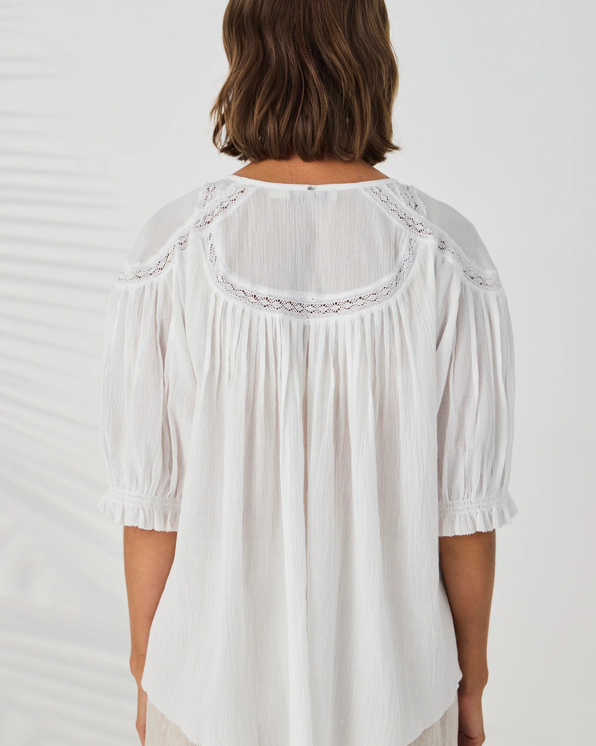 A woman viewed from the back wearing a white blouse with pleated detailing, ruffled short sleeves, and a sheer yoke with embroidered trim, paired with light beige pants against a white background.
