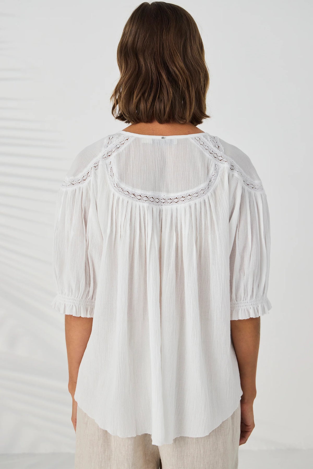 A woman viewed from the back wearing a white blouse with pleated detailing, ruffled short sleeves, and a sheer yoke with embroidered trim, paired with light beige pants against a white background.