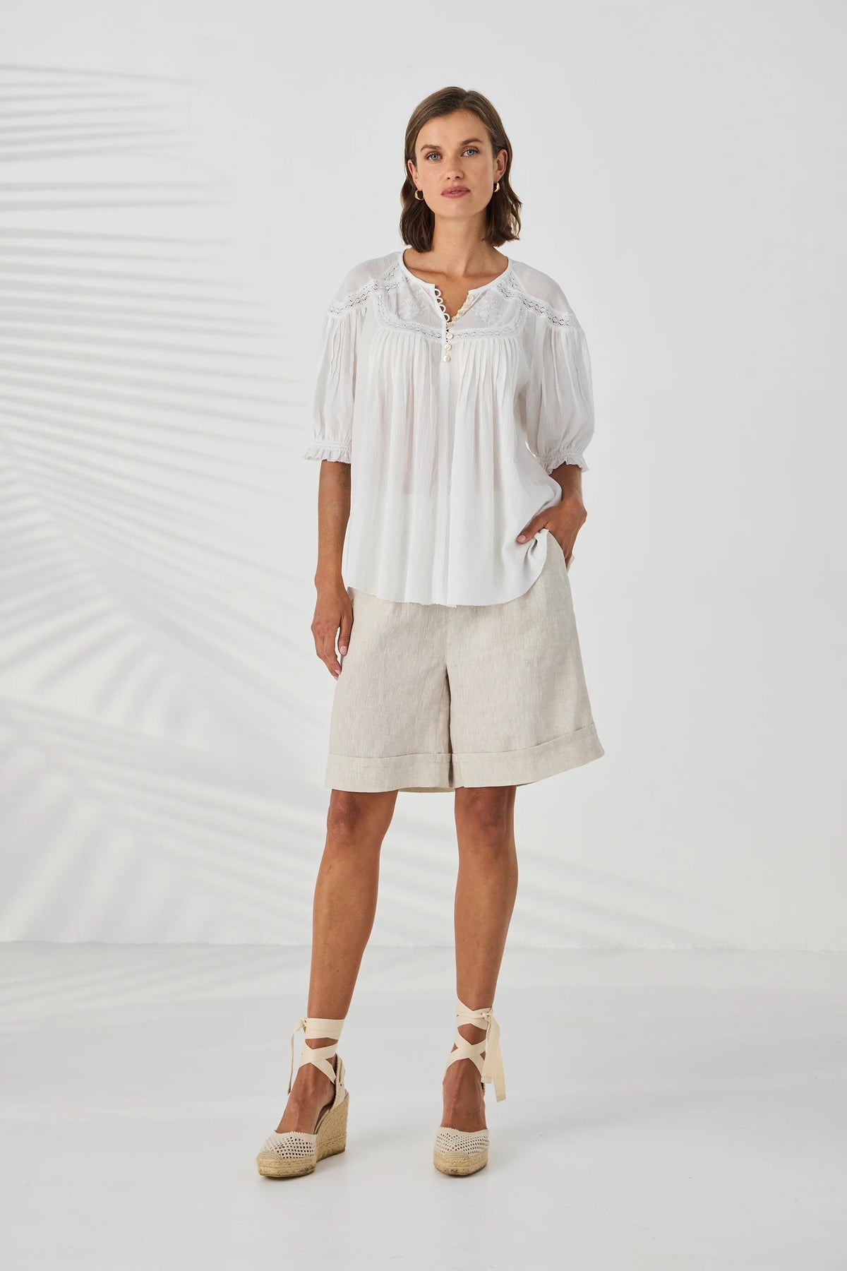 A woman modeling a white short-sleeve blouse with a split neckline and beige linen shorts against a white studio backdrop with soft leaf shadows.