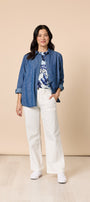 Woman wearing a blue shirt over a patterned top with white pants against a beige background