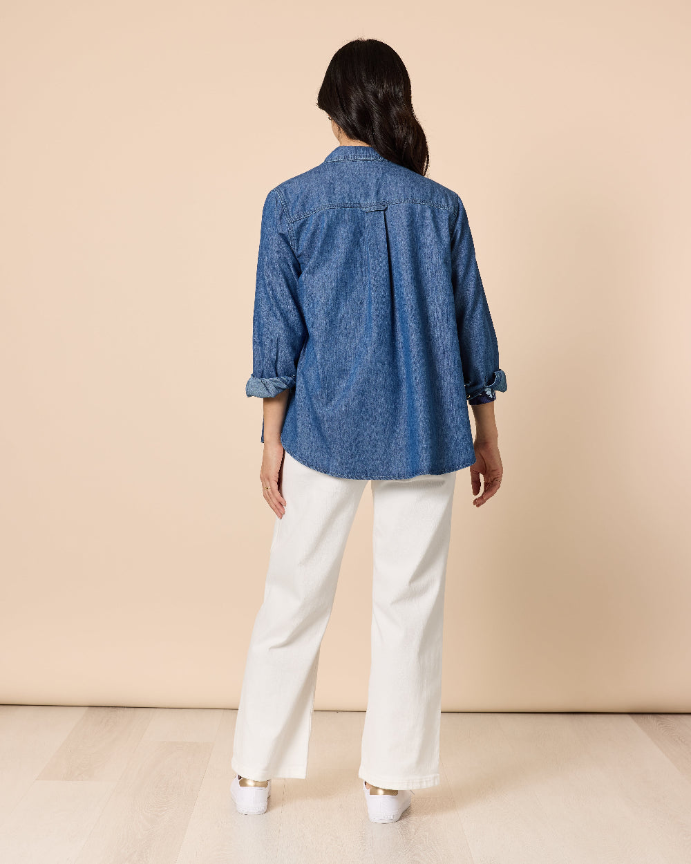 Person wearing a blue denim shirt and white pants against a beige background