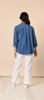 Person wearing a blue denim shirt and white pants against a beige background