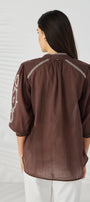  back view wearing a brown semi sheer blouse with white embroidered details on the shoulders and sleeves, paired with white pants.