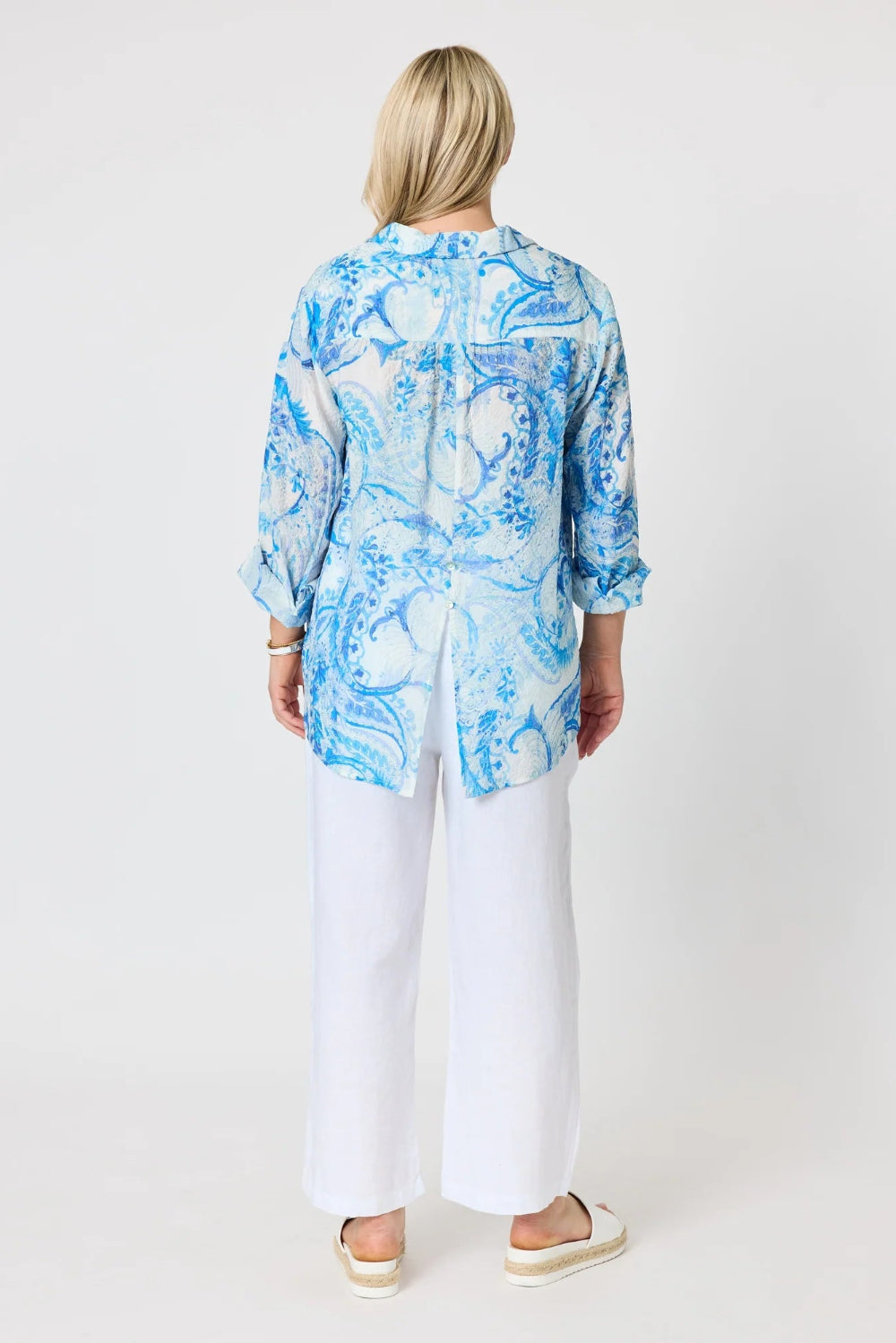 A woman viewed from the back wearing a light blue and white abstract print long sleeve blouse with white wide leg pants and white espadrille sandals.