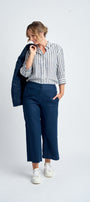 A woman wearing navy blue cropped linen pants and a striped shirt, standing against a white background, with her back to the camera. She is also wearing white sneakers.