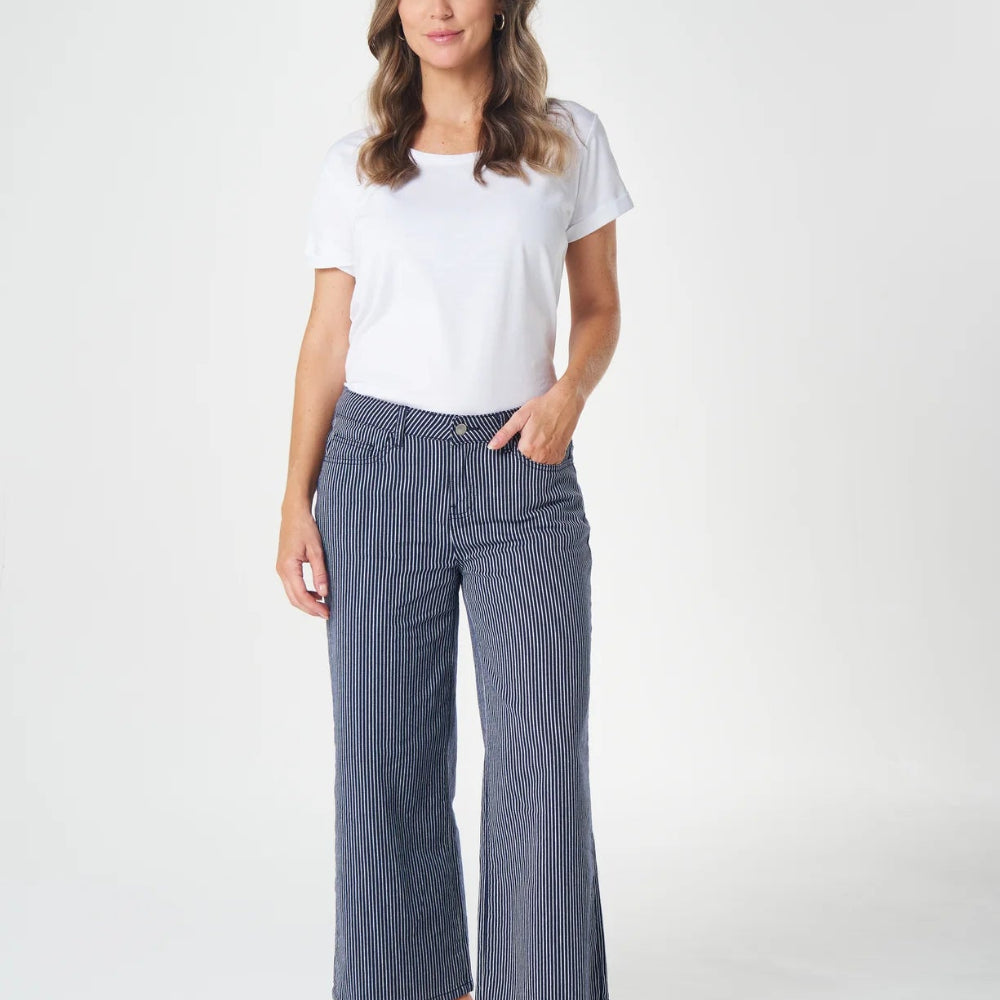 A woman modeling a pair of wide-leg, cropped blue pinstripe pants with a white t-shirt and white slide sandals, standing against a light gray studio background.