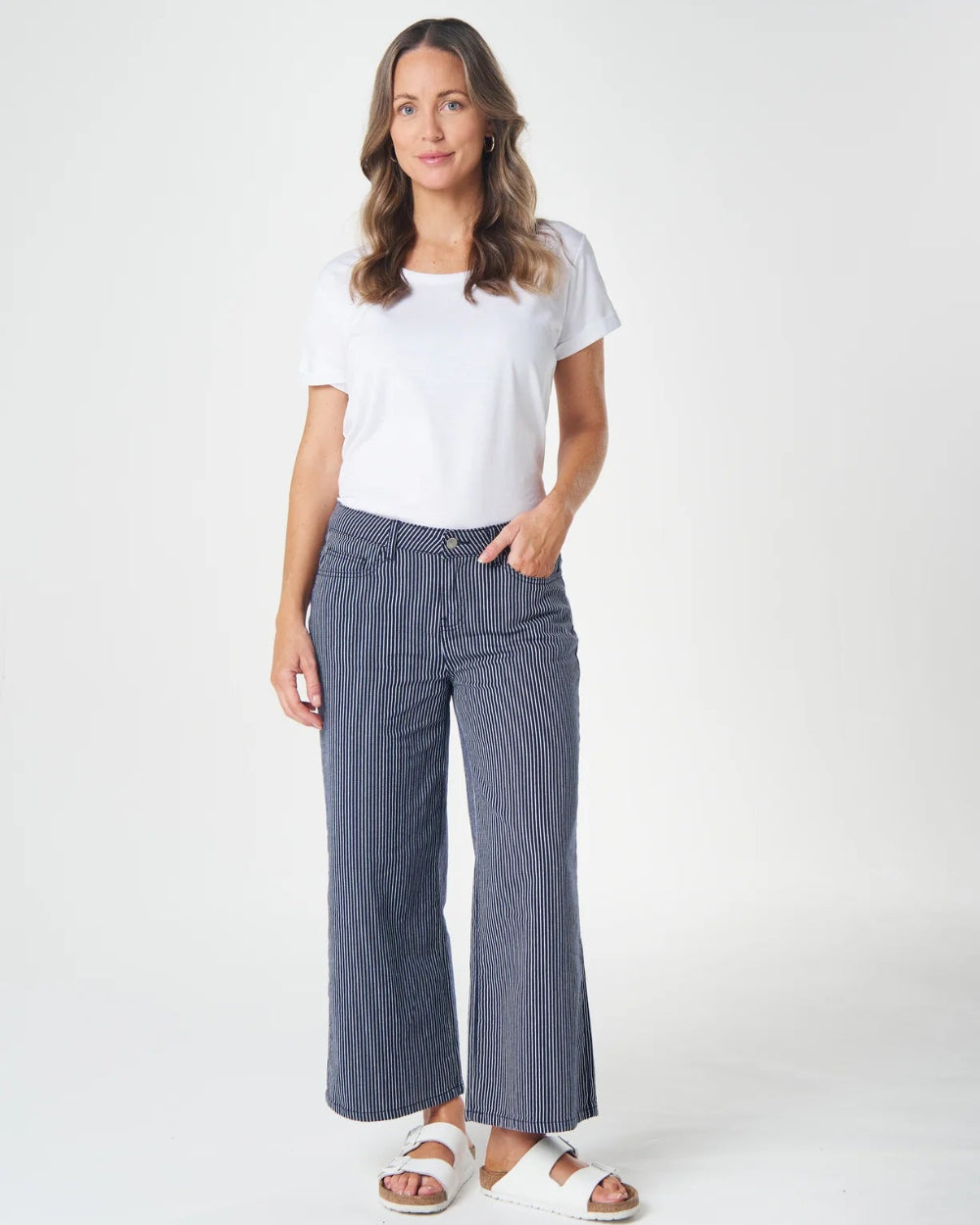 A woman modeling a pair of wide-leg, cropped blue pinstripe pants with a white t-shirt and white slide sandals, standing against a light gray studio background.