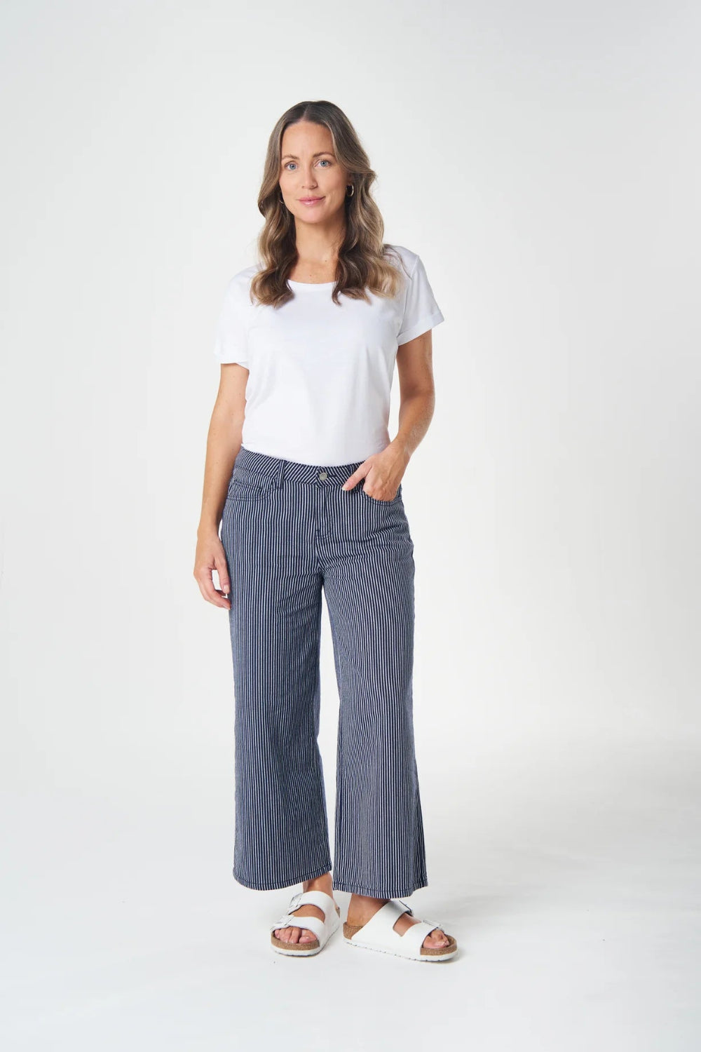 A woman modeling a pair of wide-leg, cropped blue pinstripe pants with a white t-shirt and white slide sandals, standing against a light gray studio background.