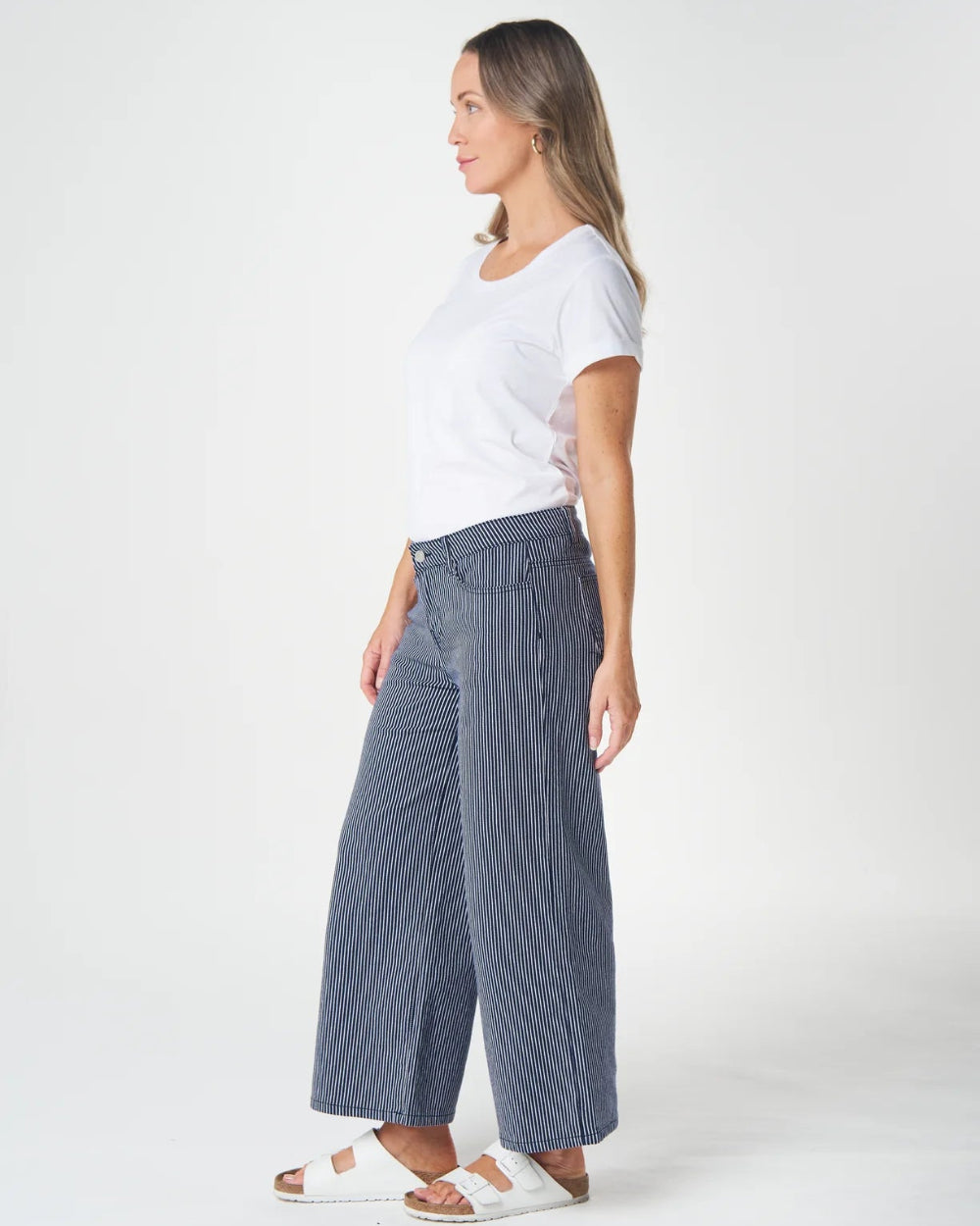 A woman shown in profile wearing a white short-sleeve T-shirt, wide-leg blue jeans, and white slide sandals against a clean white studio background.